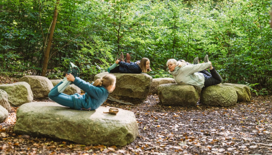 Kinderen en een volwassene liggen op grote stenen in het bos en doen samen yoga-oefeningen, omringd door dichte bomen en bladeren.