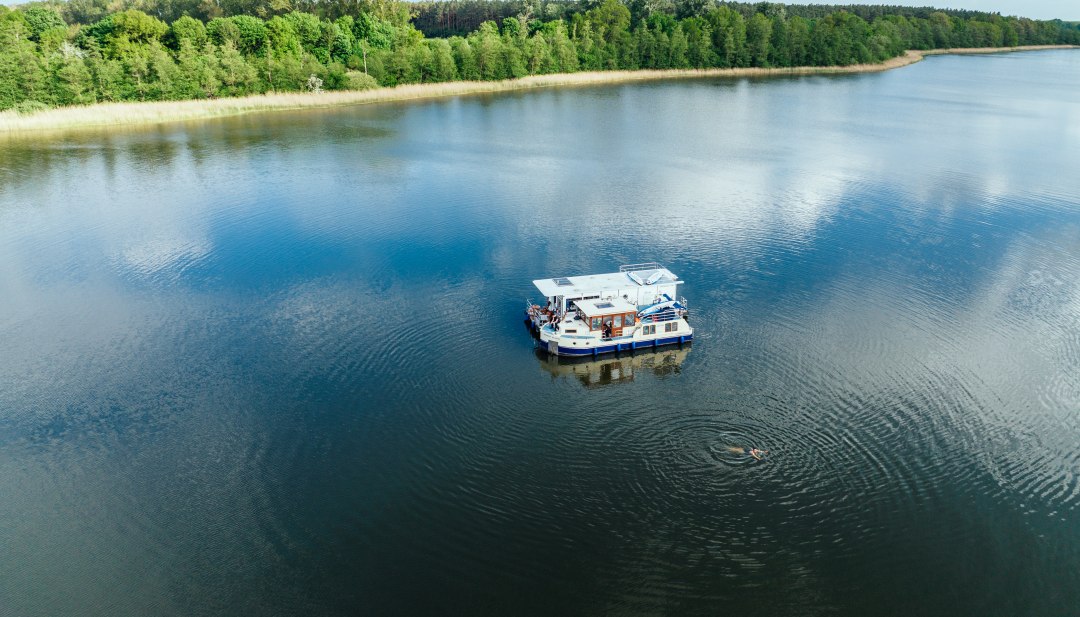 Dwie łodzie mieszkalne leżą obok siebie na jeziorze, a obok nich pływa człowiek. // Podróż łodzią mieszkalną po Pojezierzu Meklemburskim // © MV-T/Gänsicke Dwie łodzie mieszkalne leżą obok siebie na jeziorze, a obok nich pływa człowiek.