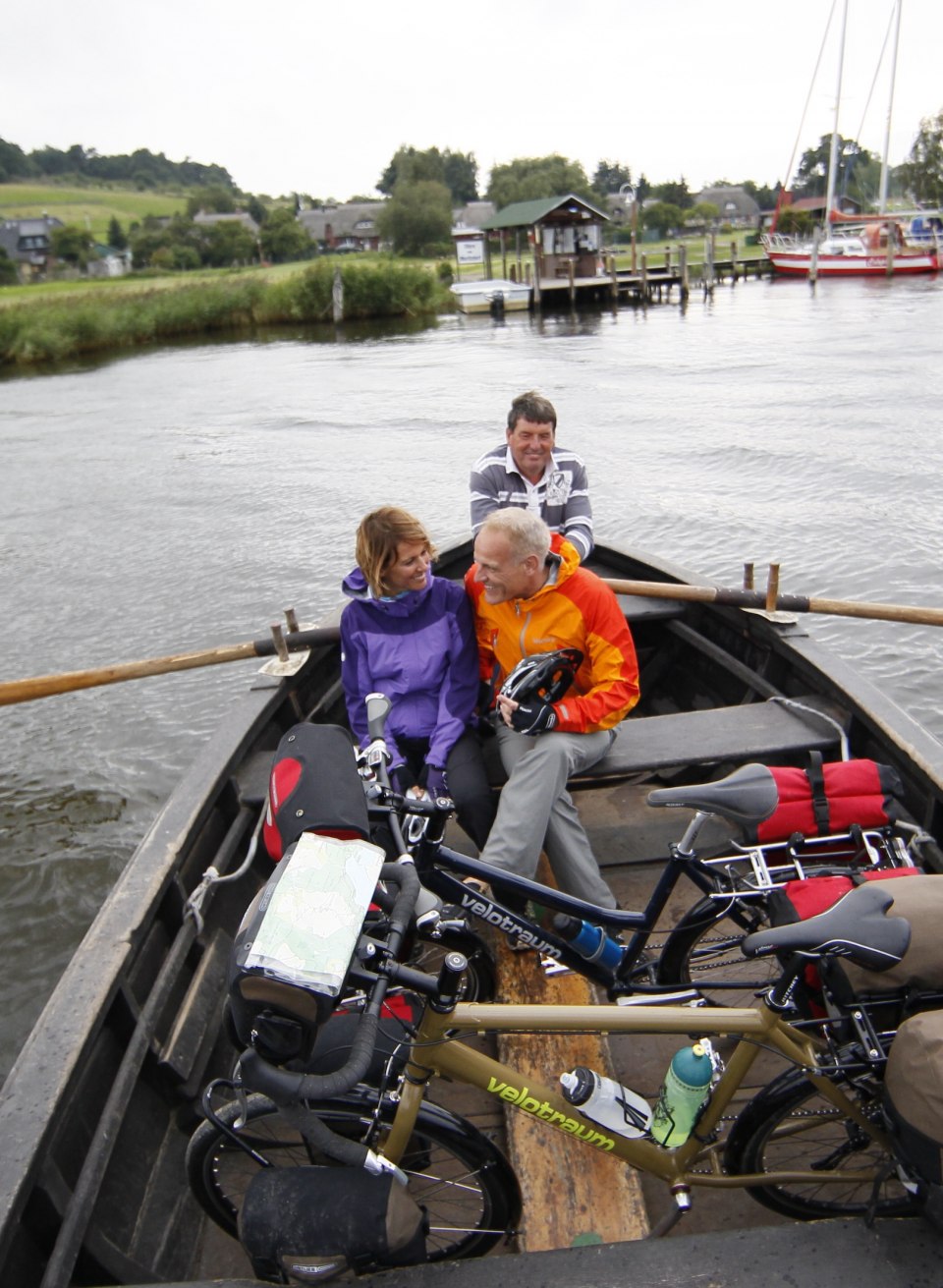 Een idyllische oversteek: fietsers gebruiken de traditionele roeipont bij het Baaber Bollwerk op Rügen om de Bodden over te steken en te genieten van de schoonheid van de natuur., © TMV/outdoor-visions.com Drie mensen met fietsen op een roeipont bij het Baaber Bollwerk, omgeven door water, met uitzicht op groene oevers en een haventje op de achtergrond.