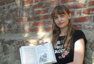 Jose Saefkow sits in a black shirt in front of an old brick wall and shows her book // &copy; Josi Saefkow