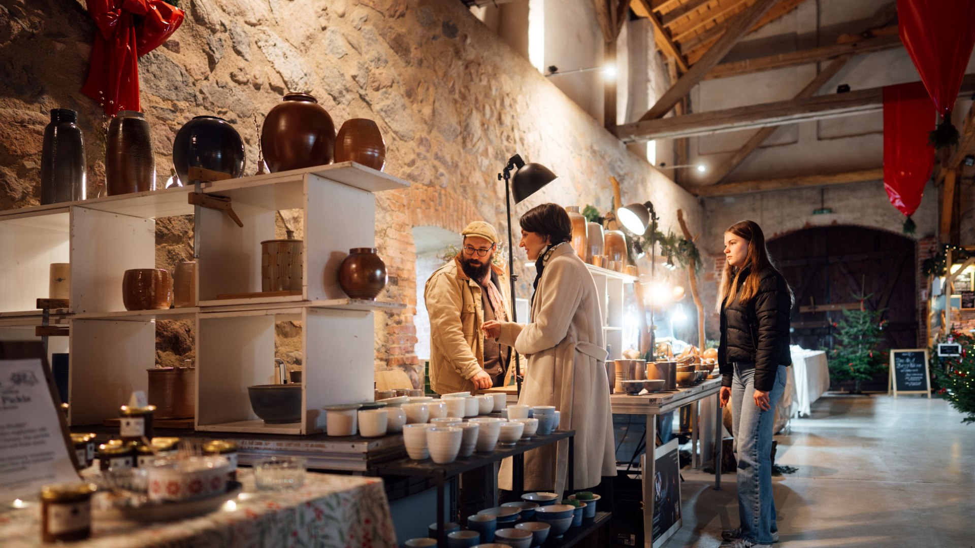 A vendor at a Christmas market presents handmade ceramic bowls and vases, while a visitor asks questions with interest.