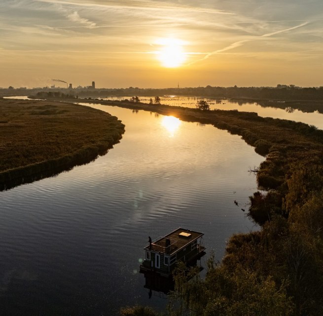 Zonsondergang over de Peene, terwijl een woonboot vredig op de rivier drijft. Het landschap strekt zich uit tot ver in de verte, waar hemel en water elkaar ontmoeten., © TMV Woonboot op de Peene bij zonsondergang, omgeven door een vredig rivierlandschap en gouden licht aan de horizon.