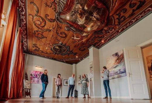 Gasten staan in de barokke hal en kijken naar het rijk beschilderde plafond met ornamenten in het landhuis van Ludorf. // Plafond omhoog, uitzicht naar boven: In de barokke hal van het landhuis Ludorf vertellen ornamenten over kunst en tijd. // © MV-T/Petermann Gasten staan in de barokke hal en kijken naar het rijk beschilderde plafond met ornamenten in het landhuis van Ludorf.