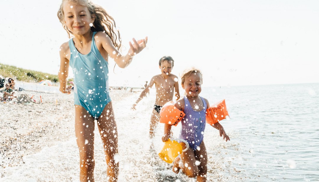 Trójka dzieci biegnie przez płytką wodę na bałtyckiej plaży, śmiejąc się i radośnie pluskając w falach.