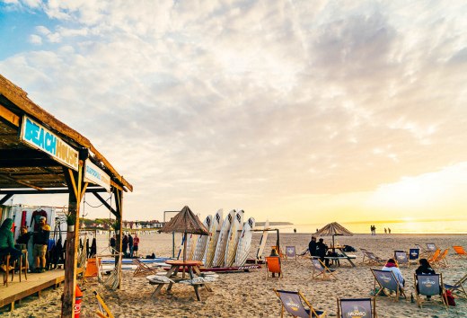 Strandsc&egrave;ne met strandhuis, surfplanken en zonsondergang in Warnem&uuml;nde, mensen ontspannen in ligstoelen.