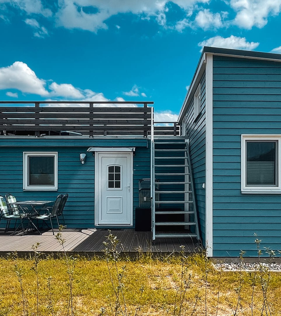Modern, blauw Tiny House met dakterras en gezellig houten terras met zitje, omgeven door landelijk landschap onder een blauwe hemel.