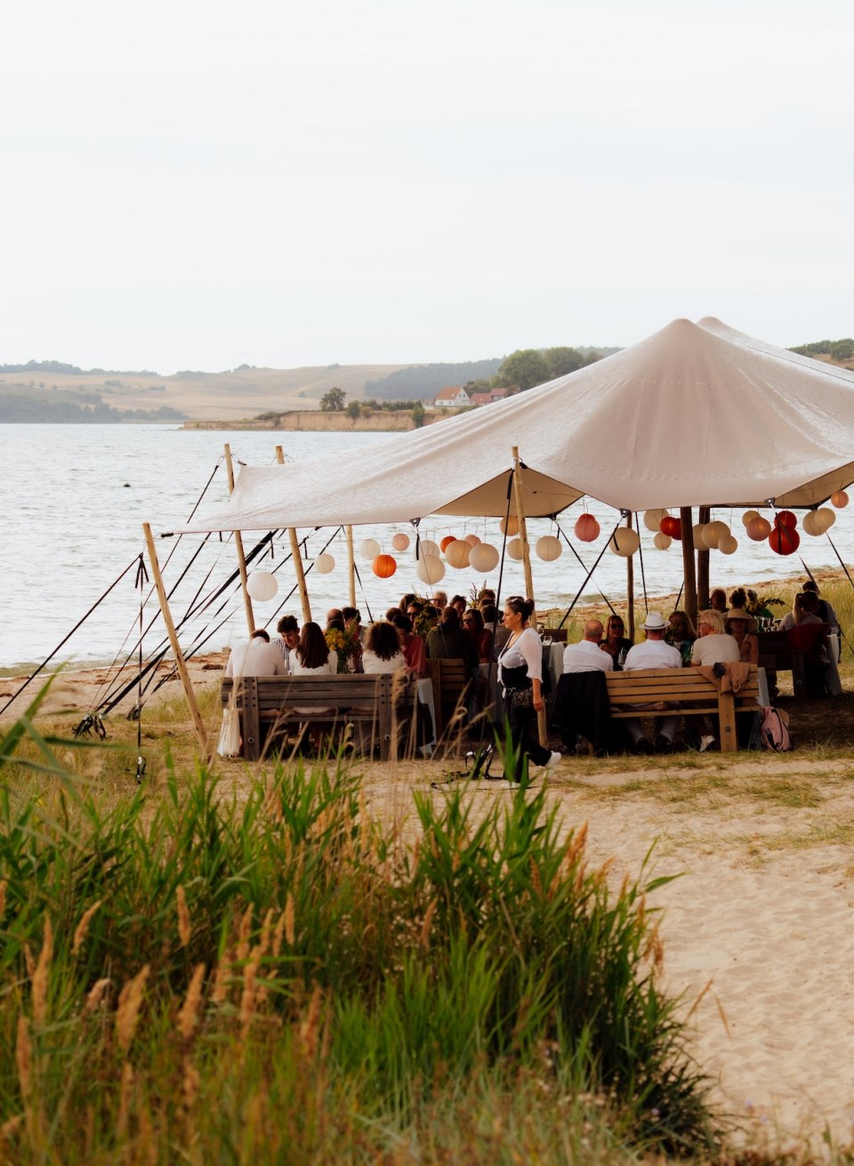 Goście siedzą przy długich stołach pod namiotem wśród trawy wydmowej na plaży w Thiessow, a tuż obok rozciąga się morze. // Pod dachem namiotu, wśród trawy na wydmach, zbiera się grupa – kolacja na plaży w Thiessow, z widokiem na wodę. // © MV-T/Petermann Goście siedzą przy długich stołach pod namiotem wśród trawy wydmowej na plaży w Thiessow, a tuż obok rozciąga się morze.