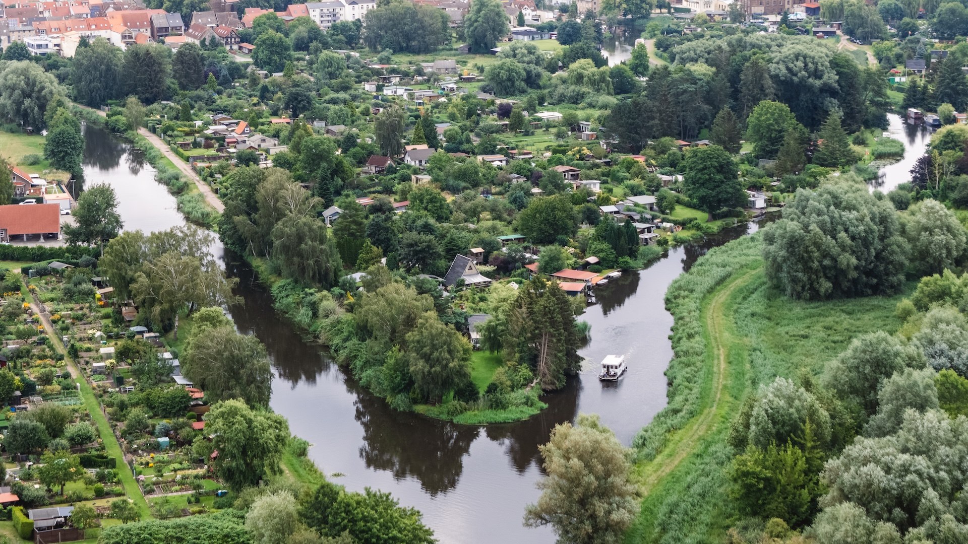 Een luchtfoto toont de kronkelende Elde met een woonboot omgeven door groene tuinen en de stad Parchim op de achtergrond.