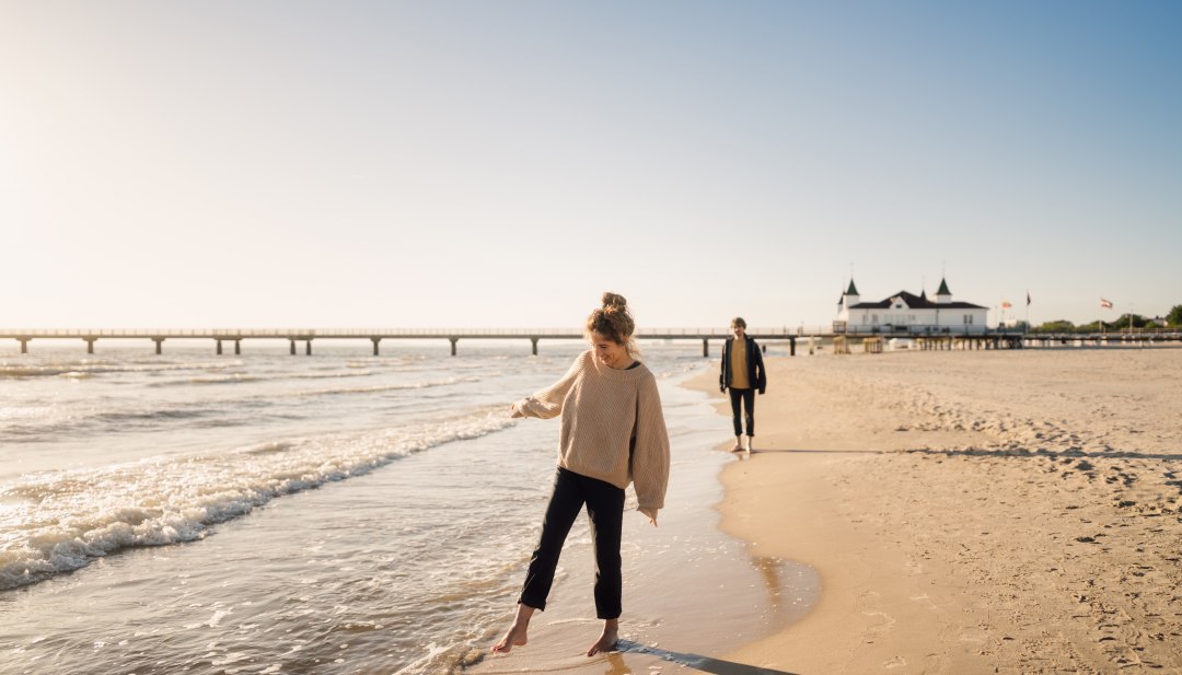 Dwie osoby cieszą się spacerem po plaży w promieniach słońca na molo Ahlbeck na wyspie Uznam. // Boso przez wodę - relaksujący spacer wzdłuż plaży przy historycznym molo w Ahlbeck na Uznamie w łagodnym wieczornym świetle. // © MV-T/Petermann Dwie osoby cieszą się spacerem po plaży w promieniach słońca na molo Ahlbeck na wyspie Uznam.