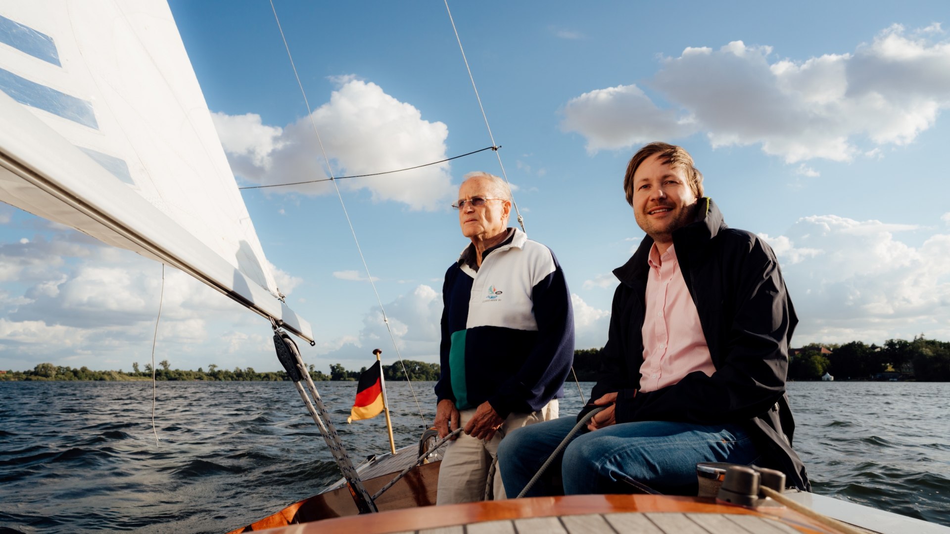 Jannis and Manfred Achtenhagen are sitting on a sailboat on Lake Müritz, with the sail and a cloudy sky in the background. // Wind in the sails, open waters ahead: Jannis Achtenhagen sits on board with Manfred Achtenhagen and lets the Müritz do its work—a clear course // © MV-T/Petermann Jannis and Manfred Achtenhagen are sitting on a sailboat on Lake Müritz, with the sail and a cloudy sky in the background.