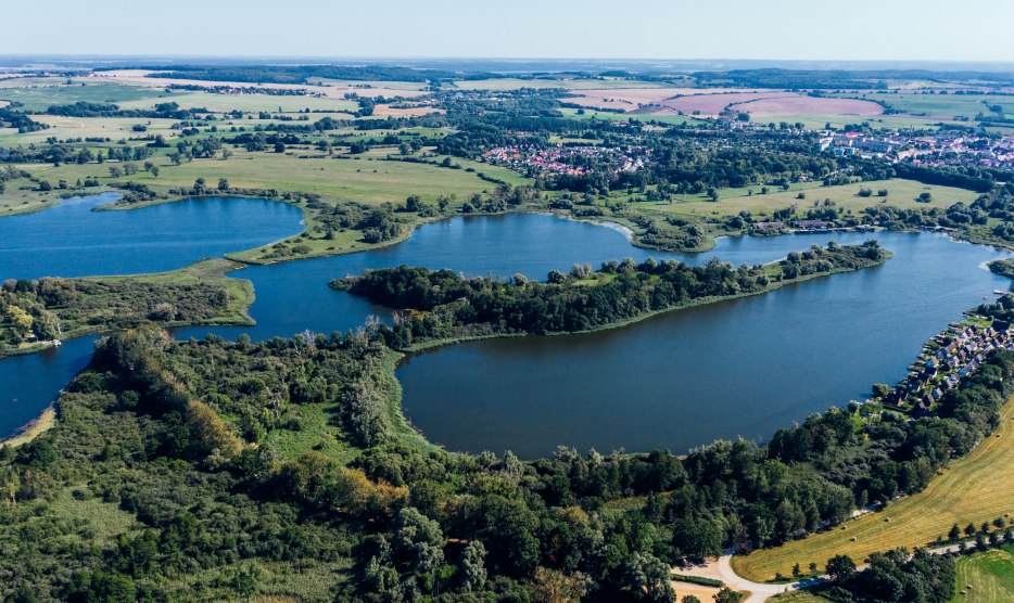 Aerial view of Lake Teterow with Burgwallinsel, green shores, fields and Teterow in the background.