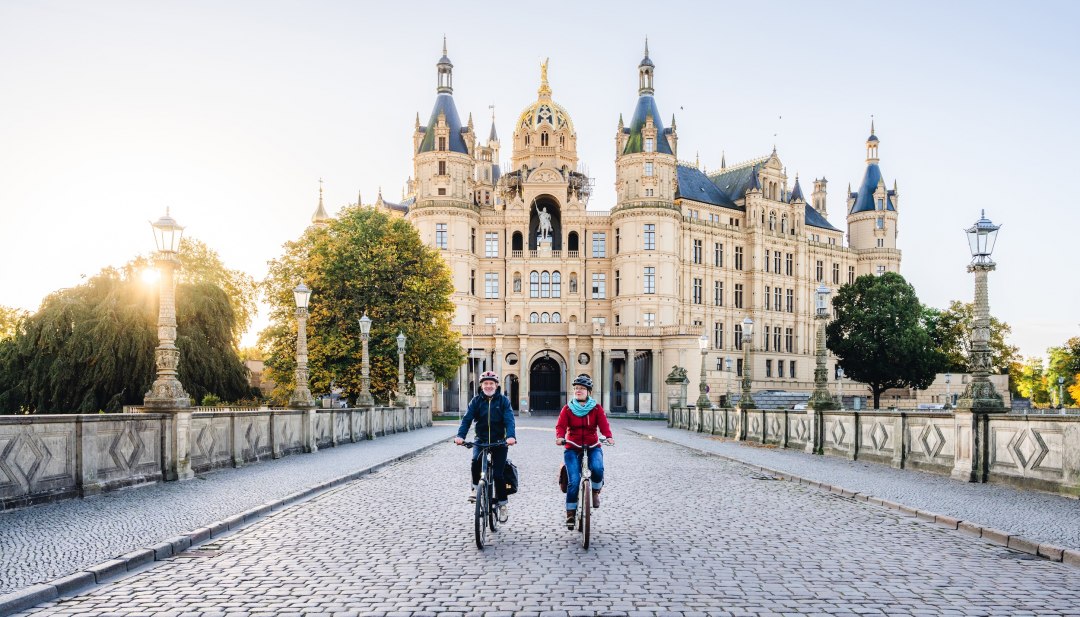 Dwóch rowerzystów przejeżdża przez most przed zamkiem w Schwerinie, oświetlonym złotym porannym słońcem. // Bajkowe poranne słońce - Zamek Schwerin w jesiennej scenerii zaprasza na wycieczkę rowerową. // © MV-T/Gross Dwóch rowerzystów przejeżdża przez most przed zamkiem w Schwerinie, oświetlonym złotym porannym słońcem.