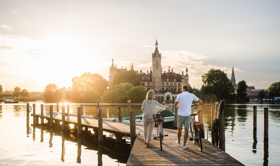 A couple with bicycles stand on a jetty on Lake Schwerin and look at Schwerin Castle in the evening light.