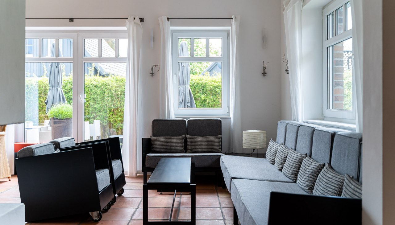 A bright and stylishly furnished lounge area with large windows that let in plenty of natural light. Gray sofas and armchairs with minimalist black accents invite you to relax. The warm terracotta floors and simple white curtains give the room a relaxed and inviting atmosphere., © Hotel Carpe Diem