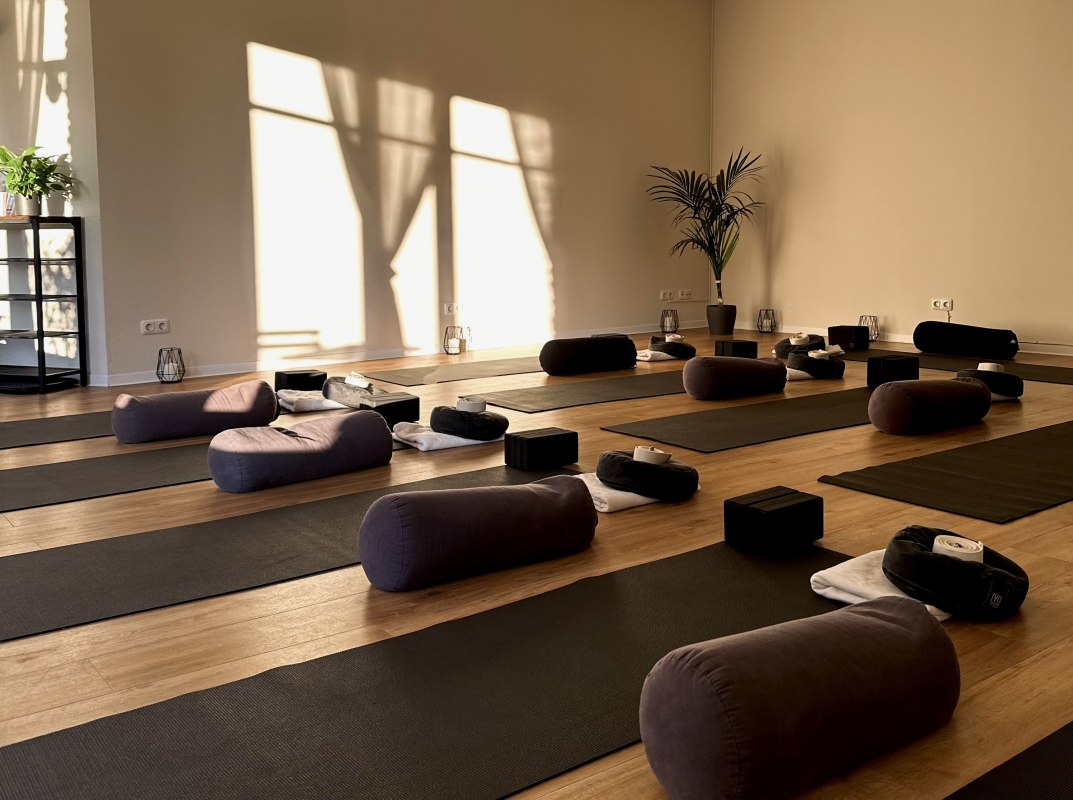Bright, minimalist yoga room with wooden flooring, several yoga mats, meditation cushions, bolsters and yoga blocks. Warm sunlight shines through large windows and creates a calm, relaxing atmosphere for yoga, meditation and relaxation., &copy; MOVA PLACE - Yoga & Co. Studio