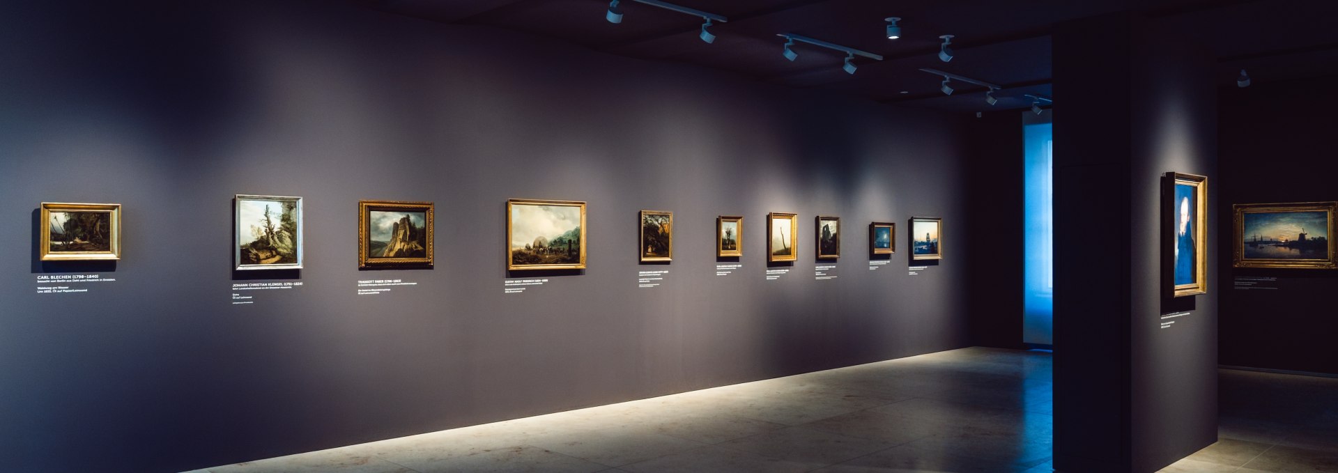 Epoka romantyzmu rozwija się w nastrojowo oświetlonych salach Pomorskiego Muzeum Państwowego w Greifswaldzie. Arcydzieła Caspara Davida Friedricha i jemu współczesnych zapraszają zwiedzających do spokojnego dialogu ze sztuką. Każdy obraz otwiera okno do minionych światów pełnych tęsknoty i bliskości z naturą., © MV-T/Gross Ciemna sala wystawowa z romantycznymi obrazami na ciemnoszarej ścianie w Pomorskim Muzeum Państwowym w Greifswaldzie.