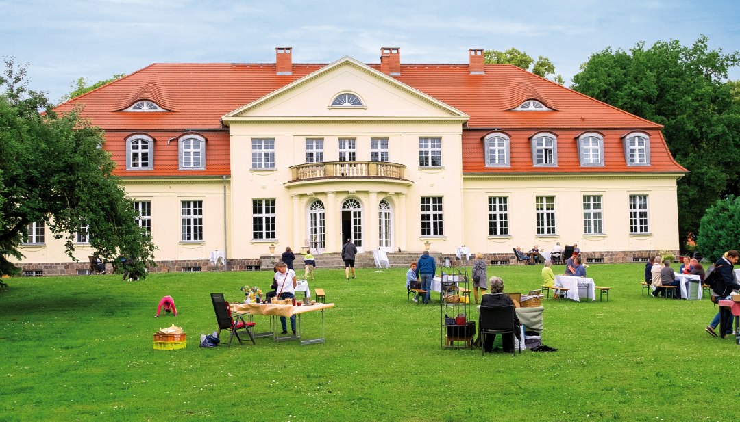 Mensen zitten op het gazon voor het landhuis Gevezin tijdens een midzomerevenement, terwijl het historische gebouw schittert in het daglicht.