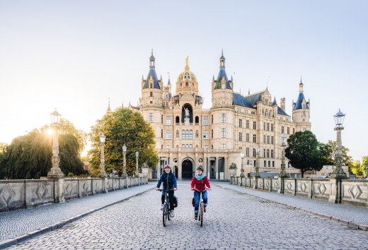 Dwóch rowerzystów przejeżdża przez most przed zamkiem w Schwerinie, oświetlonym złotym porannym słońcem.
