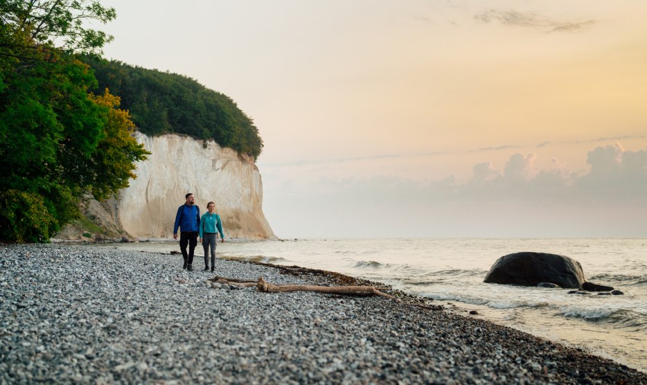 Een koppel wandelt langs de voet van de beroemde krijtrotsen op het eiland Rügen, terwijl de zonsondergang de hemel in zachte kleuren oplicht.