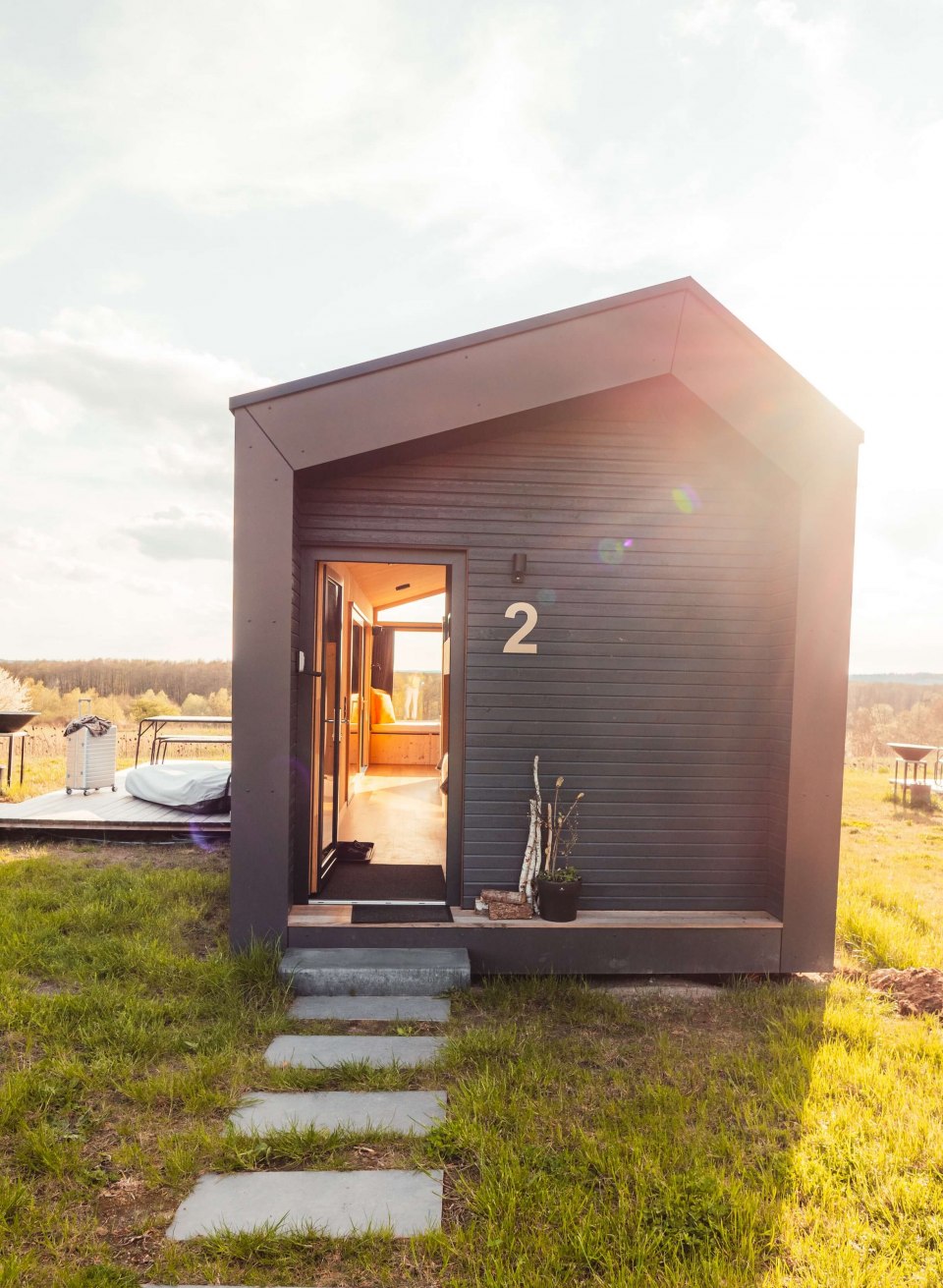 Klein, donker huisje met houten lambrisering, open ingangsdeur en zonnestralen op een groene weide.