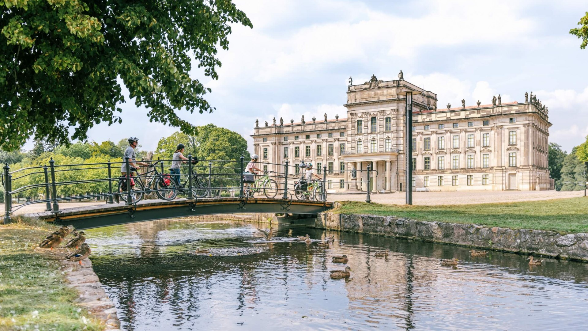 Kasteel Ludwigslust met fietsers die het kanaal oversteken op een brug. // "Versailles van het Oosten' en 'Sanssouci van het Noorden' - Paleis Ludwigslust is een absoluut hoogtepunt van de tour. // © MV-T/Tiemann Kasteel Ludwigslust met fietsers die het kanaal oversteken op een brug.