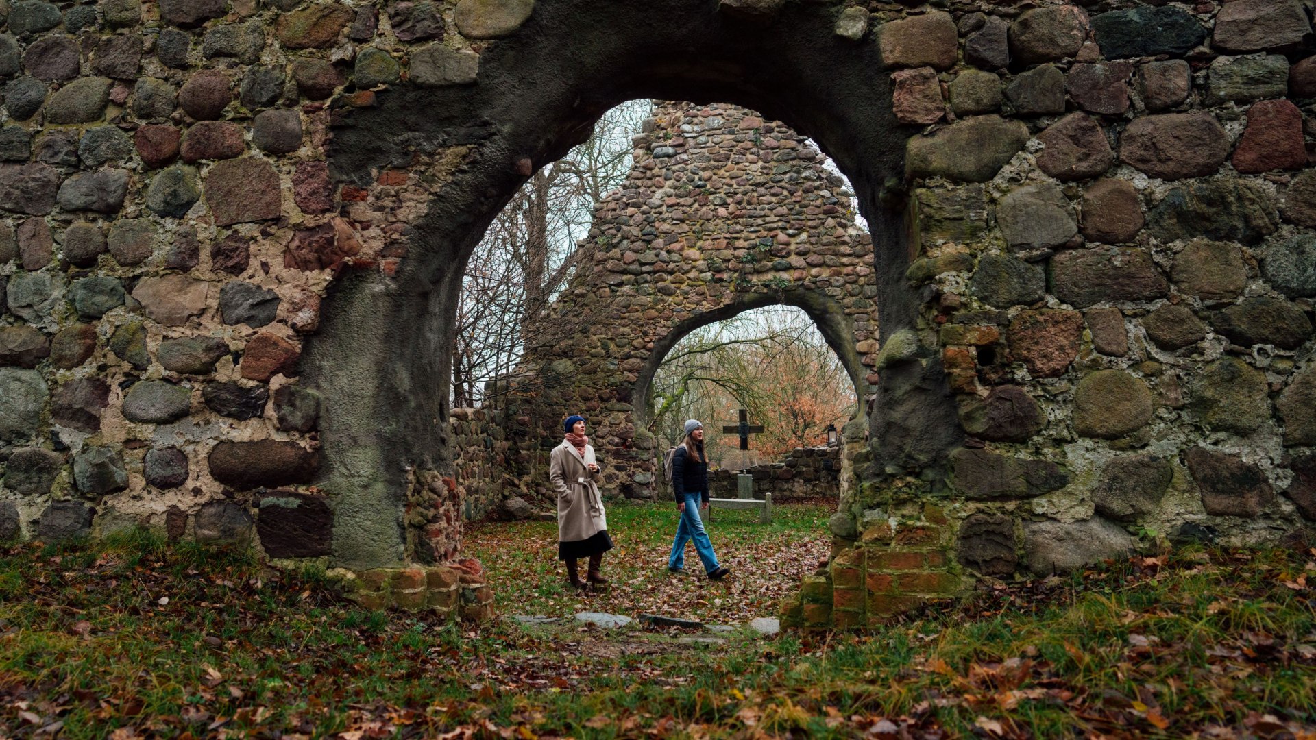Two people explore the ruined desert church in Ulrichshusen, surrounded by stone arches, autumn leaves and bare trees.