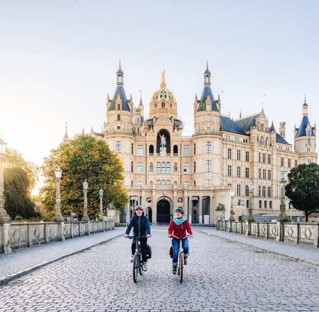 Two cyclists cross a bridge in front of Schwerin Castle, illuminated by the golden morning sun. // Fairytale morning sun - Schwerin Castle in an autumnal setting invites you to take a bike tour. // © MV-T/Gross Two cyclists cross a bridge in front of Schwerin Castle, illuminated by the golden morning sun.
