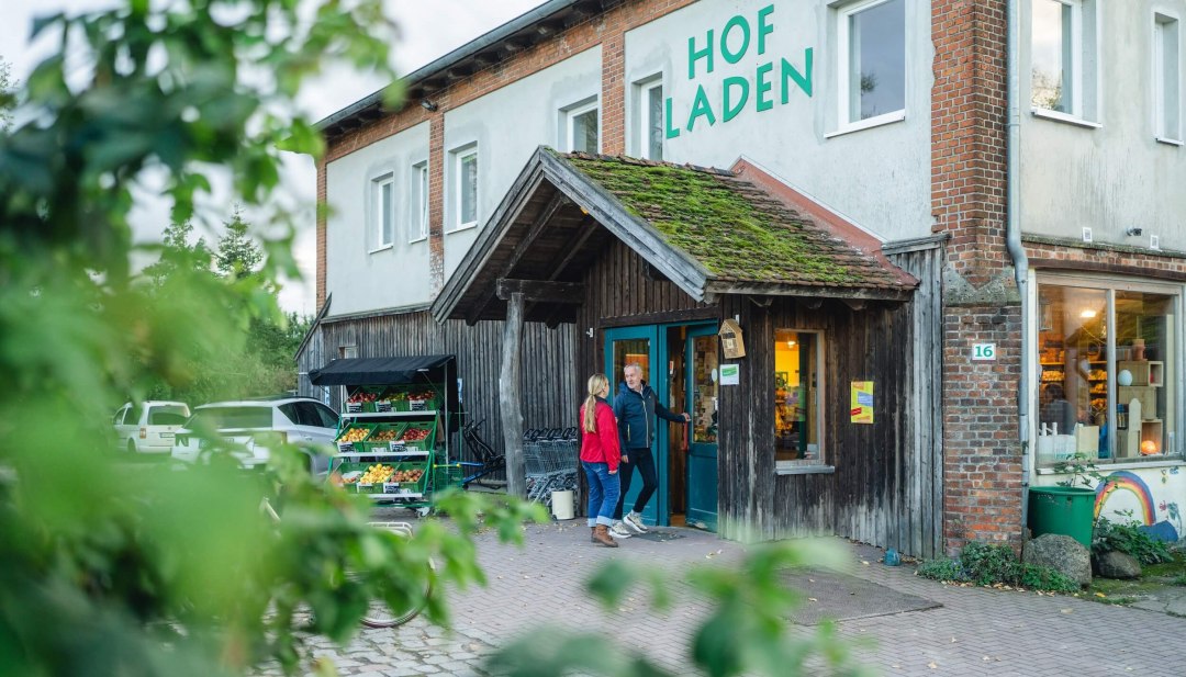 Rustic farm store with green roof, fresh fruit and vegetable stand outside and two people at the entrance.