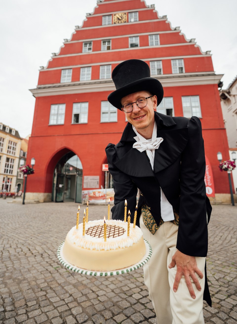 Een man met een wrak en hoed houdt een taart voor de camera op het marktplein in Greifswald ter ere van de verjaardag van Caspar David Friedrich.