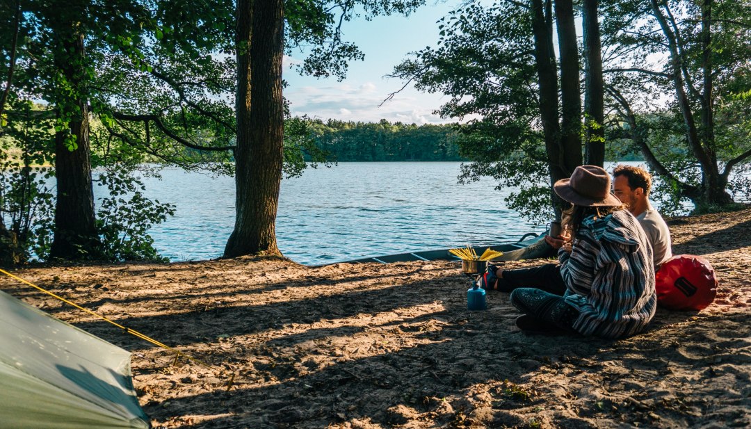 A couple sits by the water with a gas stove and cooks pasta at the Hexenw&auml;ldchen campsite