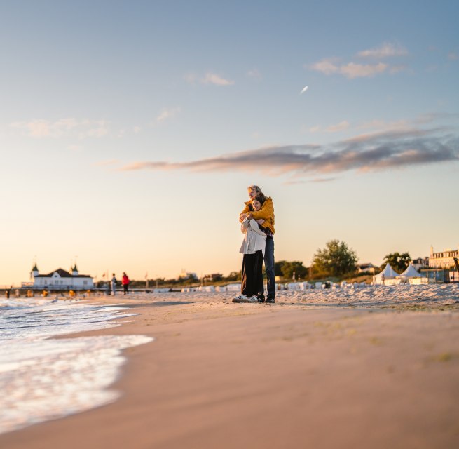 Para stoi na plaży w Ahlbeck, objęta ramionami, a molo i wieczorne niebo skąpane są w ciepłym świetle. // Zachód słońca na plaży w Ahlbeck: wspólny spacer wzdłuż brzegu, poczucie przestrzeni, chwila odprężenia. // © MV-T/Gross Para stoi na plaży w Ahlbeck, objęta ramionami, a molo i wieczorne niebo skąpane są w ciepłym świetle.