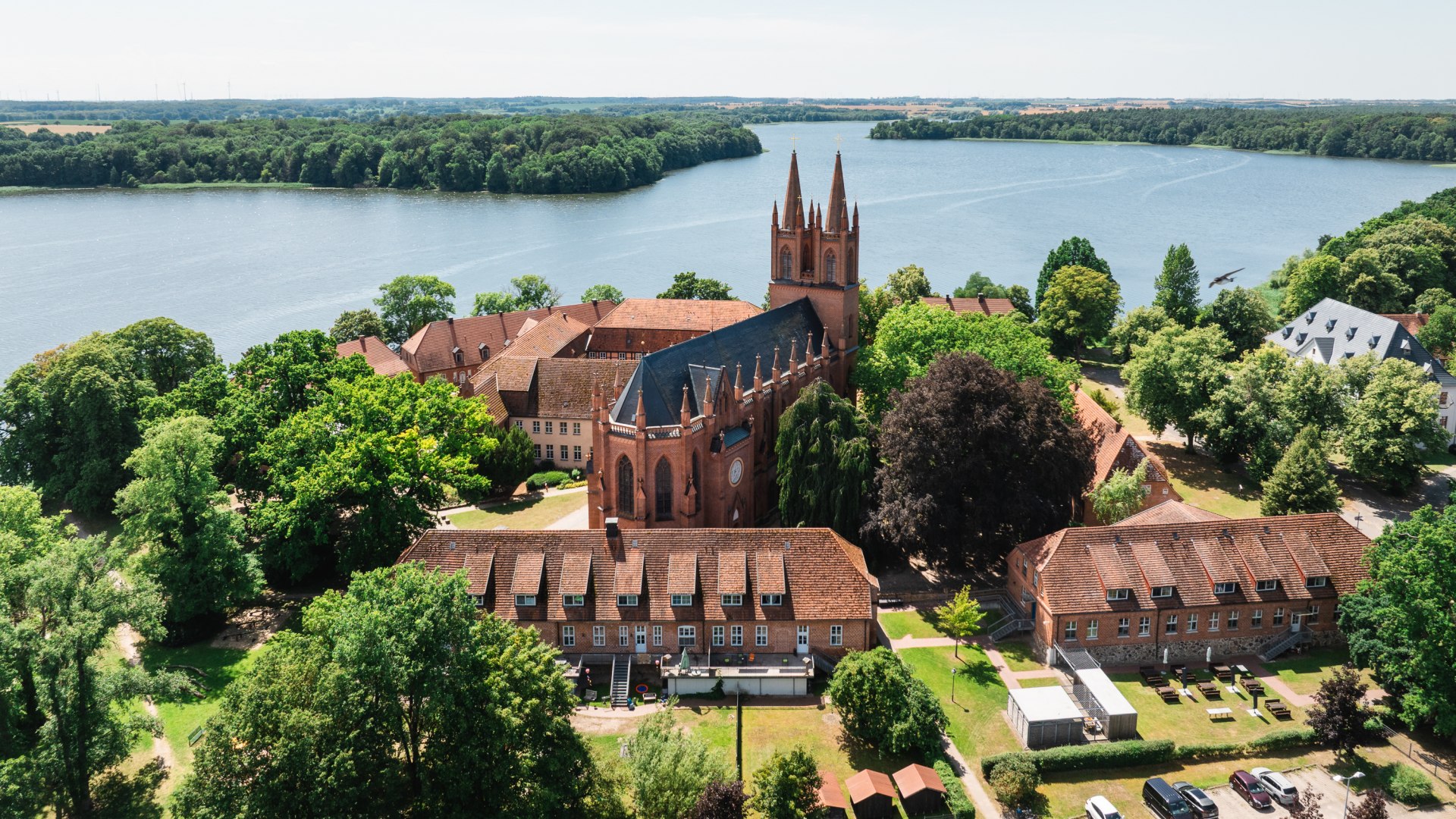 Aerial view of Dobbertin Abbey with its brick church and towers by the lake, surrounded by green trees and lakeside paths.