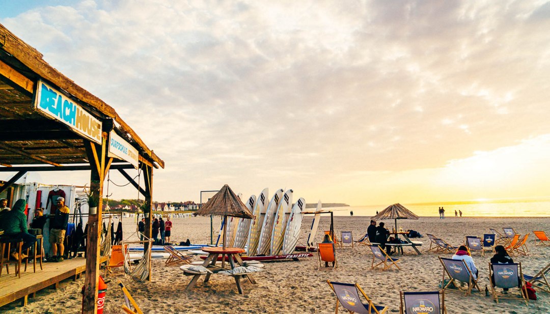 Strandscène met strandhuis, surfplanken en zonsondergang in Warnemünde, mensen ontspannen in ligstoelen.