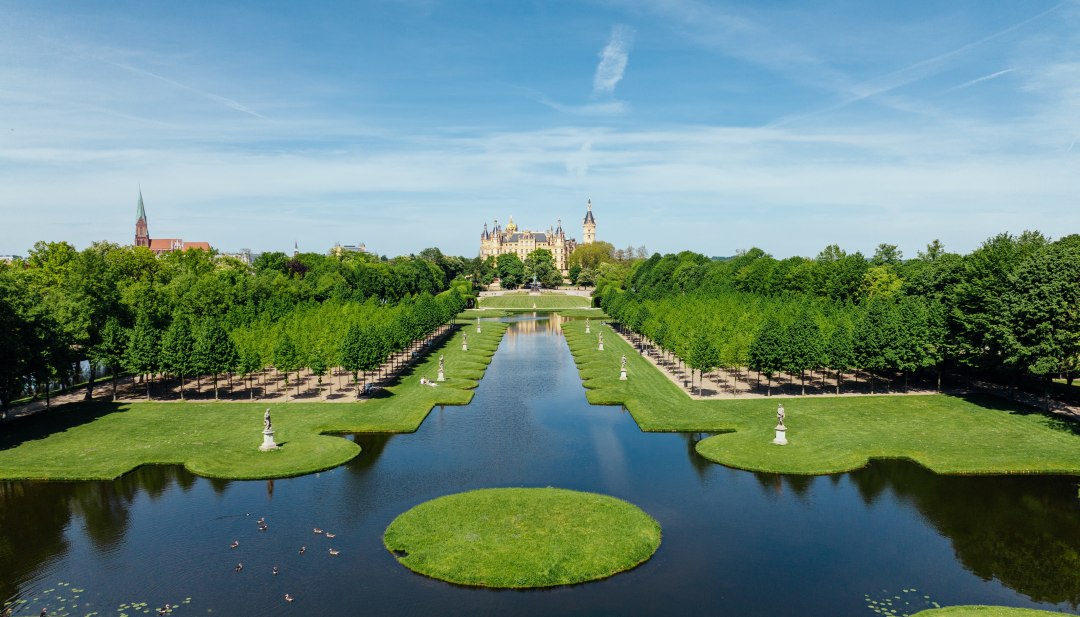 Baroktuin van Schwerin met symmetrische gracht, waterpartijen en kasteel op de achtergrond onder een blauwe hemel. // De paleistuinen van Schwerin nodigen je uit om diep adem te halen - barokke waterpartijen weerspiegelen het sprookjesachtige paleis, terwijl lanen met lindebomen voor schaduw zorgen. Ontdek waar architectuur en natuur harmonieus samensmelten. // © MV-T/Gänsicke Baroktuin van Schwerin met symmetrische gracht, waterpartijen en kasteel op de achtergrond onder een blauwe hemel.