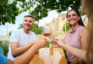 G&auml;ste sto&szlig;en beim Winzerfest Schwerin auf dem Alten Garten mit einem Glas Wein an. Im Hintergrund erhebt sich das Schweriner Schloss und gibt dem Weinfest seine besondere Kulisse. // &copy; LHSN/Celien Graubaum