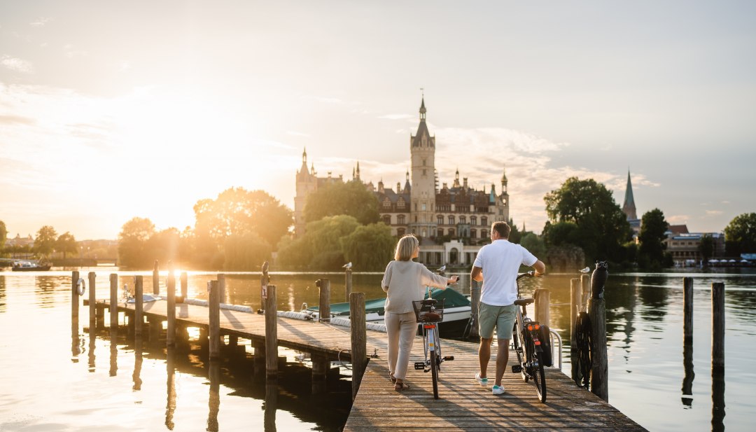 A couple with bicycles stand on a jetty on Lake Schwerin and look at Schwerin Castle in the evening light.