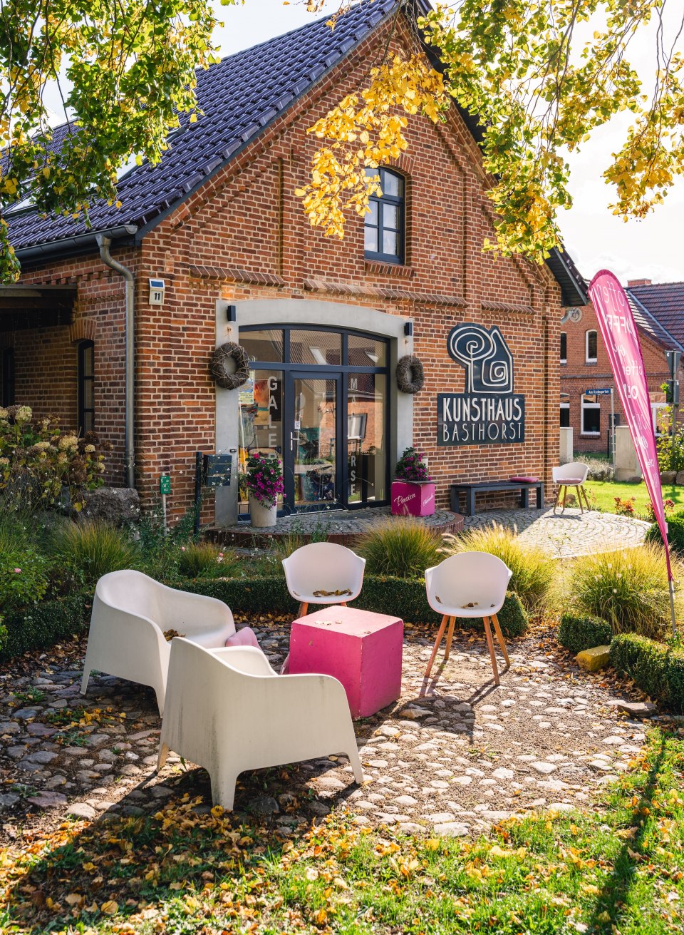 Kunsthaus Basthorst in een herfsttuin, omringd door gele en groene bladeren, met buitenzitjes.