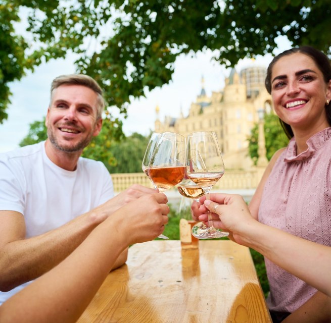 Gäste stoßen beim Winzerfest Schwerin auf dem Alten Garten mit einem Glas Wein an. Im Hintergrund erhebt sich das Schweriner Schloss und gibt dem Weinfest seine besondere Kulisse. // © LHSN/Celien Graubaum Gäste stoßen beim Winzerfest Schwerin auf dem Alten Garten mit einem Glas Wein an. Im Hintergrund erhebt sich das Schweriner Schloss und gibt dem Weinfest seine besondere Kulisse. // © LHSN/Celien Graubaum