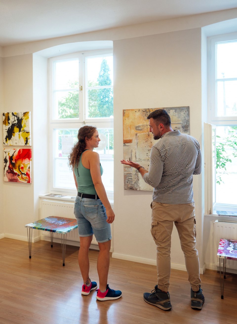 Een stel bekijkt abstracte schilderijen in de GH Abstrakt galerie in Putbus. Aan de witte muren hangen kleurrijke kunstwerken in verschillende formaten. De grote ramen laten veel daglicht binnen, wat de levendige kleuren van de schilderijen accentueert. De ruimte is licht en uitnodigend.