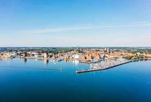 Widok z lotu ptaka na hanzeatyckie miasto Stralsund z portem, zabytkową starówką i Morzem Bałtyckim pod błękitnym niebem. // Hanzeatyckie miasto Stralsund rozciąga się między portem a zabytkową starówką. Ceglany gotyk spotyka się tu z masztami żaglowców, a wody Morza Bałtyckiego lśnią czystym błękitem. Tutaj można odkryć historię morską i tętniącą życiem kulturę portową - miejsce, które łączy w sobie wizję i przybycie. // © MV-T/Gänsicke Widok z lotu ptaka na hanzeatyckie miasto Stralsund z portem, zabytkową starówką i Morzem Bałtyckim pod błękitnym niebem.