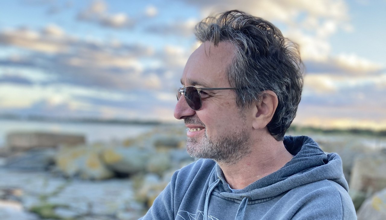 Portrait of Carl Harry Kirkeby in a blue hoodie and sunglasses in front of a rocky coastal landscape, © Carl Harry Kirkeby