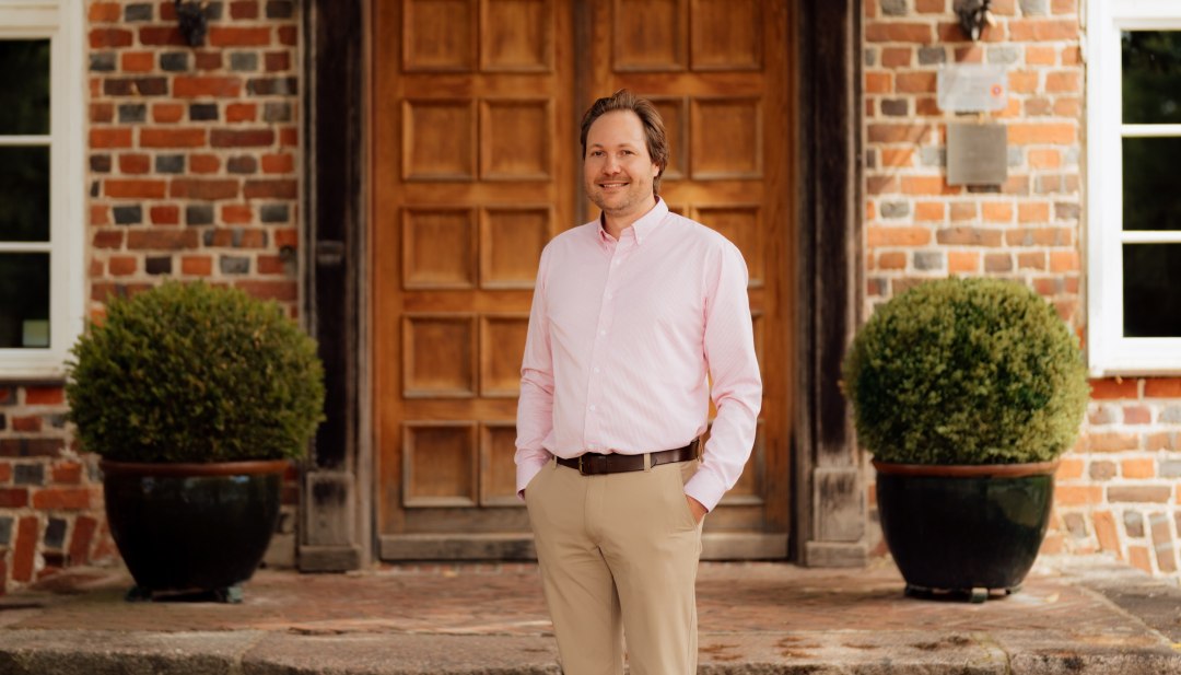 Jannis Achtenhagen is standing in front of the large wooden door of the Ludorf brick manor house, next to two round potted plants. // Jannis Achtenhagen stands in front of the historic entrance to the Ludorf manor house—a host with an eye for detail, with brickwork and a coat of arms serving as the backdrop in Mecklenburg-Western Pomerania. // © MV-T/Petermann Jannis Achtenhagen is standing in front of the large wooden door of the Ludorf brick manor house, next to two round potted plants.